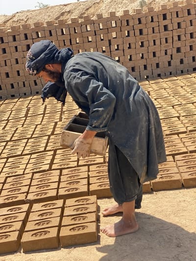 A barefoot man wearing a black head covering and dark clothing is placing bricks on the ground in Deh-E Bagh, Afghanistan. The bricks are arranged in neat rows across a dirt surface, with a stacked brick wall behind him. The man is holding a rectangular mold, presumably used to form the bricks. The scene is bathed in bright sunlight.