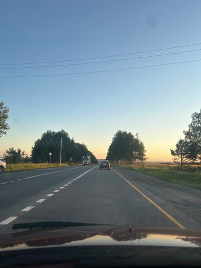 The image depicts a tranquil view from inside a vehicle traveling along a rural highway at either dawn or dusk, indicated by the warm, golden light on the horizon transitioning to a cool blue sky overhead. The scene shows an asphalt road extending into the distance, with a solid yellow line marking the left edge and a dashed white line separating the two lanes further ahead.
Several subjects are visible on the road: a dark-colored car and a white semi-truck are driving away from the camera in the right lane. On the far right, a dark, reflective surface of the vehicle from which the photo was taken is prominent. On the far left, a portion of a white car's wheel and body can be seen, possibly parked off the shoulder.
The road is flanked by dense foliage and trees on both sides, with patches of green grass visible beyond them. In the distance, a hazy landscape stretches towards the horizon. Power lines cut across the upper left section of the sky.
Notable details include two road signs on a pole to the right of the road: a red circular sign with a diagonal white line, often indicating a prohibition (such as "no entry" for certain vehicles), and below it, a rectangular white sign with a red border displaying the number "70," signifying a 70 km/h speed limit. A faint, dark circular smudge is visible in the upper left sky, likely a camera lens or sensor dust spot. The overall impression is that of a peaceful journey through the countryside near Semenovo, Russia, during a scenic time of day.