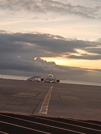 An airplane sits on an airport runway with the ocean in the background under a cloudy sky at sunrise or sunset. A boarding ramp is attached to the plane. The runway has yellow markings. The bottom of the photo shows dark, blurred lines, perhaps part of a railing or structure. The overall scene is muted and calm.
