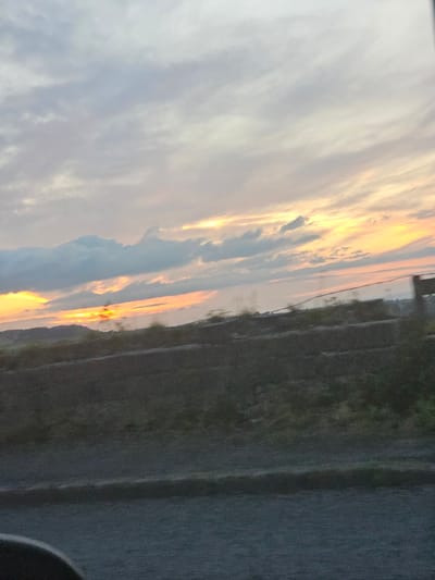 The image depicts a sunset over the horizon in Gorsedd, United Kingdom. The sky is filled with varying colors of clouds, ranging from white and gray to orange and yellow, indicating the setting sun. Below the horizon, there's a glimpse of dark land. The photograph appears to be taken at ground level. Vegetation is visible and a grey surface fills a portion of the right side of the image.