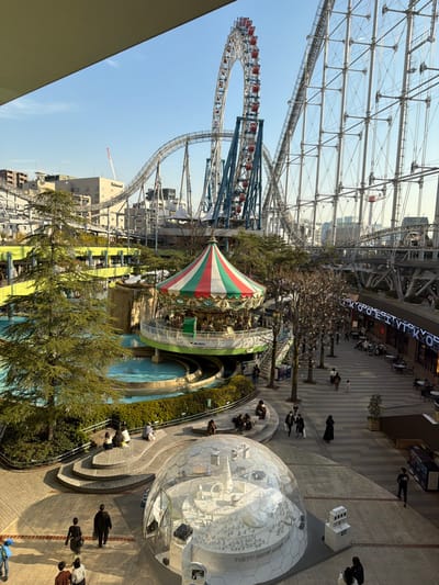An amusement park features a Ferris wheel and roller coaster under a steel lattice structure. A carousel with a striped tent-like roof is centered near a small pool. People stroll along a brick path toward a geodesic dome housing a miniature city. Buildings and trees are visible in the background.