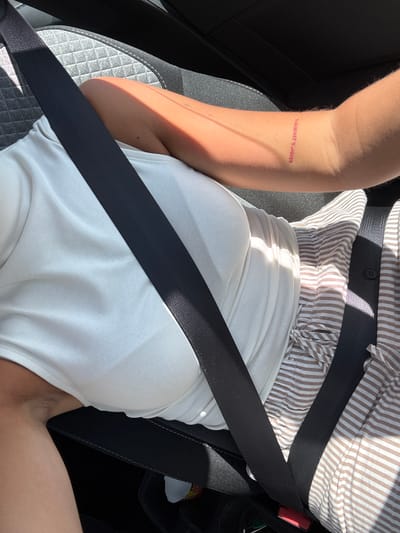 The image depicts a person wearing a white tank top and a striped shirt in a car, fastened with a seatbelt. The person has a tattoo on their left arm that reads "LOCATION'S NAME". Based on the tattoo and the context of the prompt, it's possible the location refers to Le Muy, France. The person is sitting on a gray car seat.