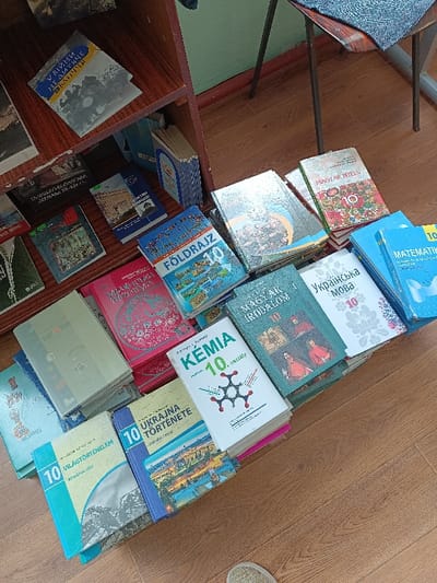 The image shows a stack of textbooks arranged on a wooden floor in a classroom. The textbooks are various colors, mostly blue and red, with white or black titles. The titles are written in Hungarian and Ukrainian. The books are likely for a 10th grade class, as many of them have "10" on the cover. The books are piled on a wooden shelf unit. The coordinates provided place the classroom in Berehove, Ukraine. The image was likely captured on a school day while the classroom was empty. A foot is visible in the bottom right of the image, suggesting someone is present in the room and took the picture. There is a wooden chair and a table, likely part of the classroom furniture, visible in the top right corner of the image.