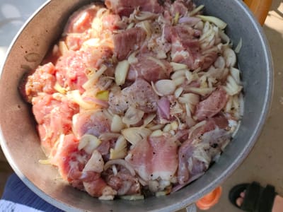 A close-up, overhead shot shows a large, grey metal pot filled with raw pork and sliced onions. The pork is cut into irregular pieces, some with visible fat. The onions are thinly sliced and mixed throughout the meat. The ingredients appear to be seasoned with black pepper. The pot is partially filled with a clear liquid, possibly marinade or oil. The lighting suggests it is daytime, with sunlight casting shadows on the left side of the pot. The bottom right corner of the image shows a black sandal with a person's foot wearing it. The scene is set outdoors, with a light brown, textured surface visible around the pot, possibly concrete or dirt. The background is slightly blurred, but hints at a simple, perhaps rural, setting. It is unclear if this scene is specifically in Florești, Moldova, as no identifying markers are present.