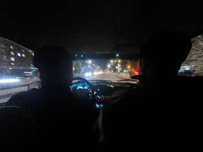 The interior of a car at night shows two people in silhouette. The driver grips the steering wheel, the dashboard illuminated. Through the windshield, streetlights line a road with sparse traffic. Buildings faintly gleam on the left. The passenger's head and shoulder are partially visible on the right. The scene is heavily underexposed.