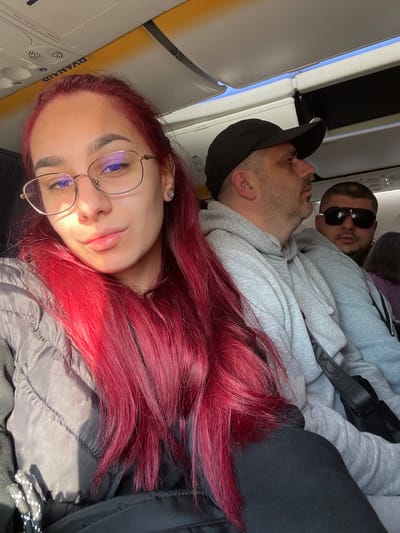 The scene depicts the interior of a Ryanair aircraft cabin, likely at or departing from Palma de Mallorca Airport (PMI) in Son Santjoan, Spain. The image features three primary individuals seated in passenger rows during daylight.

In the foreground, a female with long, bright red hair and fair complexion is captured in a self-portrait. She wears round, thin-framed eyeglasses, a small stud earring in her left ear, and a dark grey quilted jacket over a dark top. Her lips are slightly parted in a faint smile, and her gaze is directed towards the camera. Bright sunlight illuminates her left side, casting shadows on her right.

Behind and to the right of the female, a Caucasian male is seated. He wears a black baseball cap, a light grey hooded sweatshirt, and has short facial hair. He is looking forward and slightly to the right, not directly engaging with the camera. A black shoulder strap is visible across his chest.

Further back and to the right, a third Caucasian male is partially visible. He has a dark, short beard and is wearing dark sunglasses and a dark top. A prominent silver hoop earring is visible in his right ear. His head is turned, and he appears to be looking towards the camera. A small portion of a fourth individual's dark hair is visible behind him.

The aircraft cabin features overhead luggage compartments, one of which displays a "RYANAIR" decal. The ceiling and side paneling are light-colored. A window to the right reveals a bright blue sky.