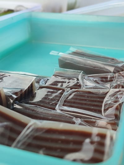 The image captures a close-up view of several individually wrapped, layered snacks, likely traditional Indonesian "kue lapis," presented in a light blue or turquoise plastic container. Each snack is rectangular and features distinct alternating layers of dark brown (resembling chocolate or palm sugar) and a lighter, off-white or cream color. They are individually sealed in clear, transparent plastic packaging, which shows some reflections of ambient light and subtle indications of moisture or small air bubbles.
The snacks are neatly arranged in rows within the container, filling a significant portion of the frame. The foreground shows several pieces in clear focus, revealing the intricate layering, while pieces further back are slightly out of focus, creating a sense of depth. The background is blurred, showing indistinct light-colored elements that could be another container or a surface.
The bright, diffused lighting suggests the photograph was taken during the daytime. There are no people visible, and no discernible text appears on any of the packaging or the container. The scene depicts a prepared display of these sweet, layered treats, often found in Indonesian markets or celebrations, aligning with the given location context of Pulorayeuk, Indonesia.