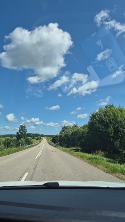 The image shows a view from inside a vehicle traveling on a paved road in Lučķini, Latvia. The road is lined with green grass and trees on both sides, and extends into the distance, curving slightly to the left. The sky is blue with scattered white clouds. The dashboard of the vehicle is visible in the foreground.