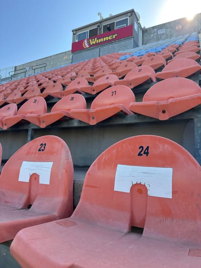 The image shows a section of stadium seating, likely at the Ness Ziona Stadium in Ness Ziona, Israel. The seats are orange and numbered, ranging from 23 to 27. Some seats have a white label covering a portion of the seat. A building with a "Winner" sign is visible in the background, with some blue seats. The scene is outdoors, with a bright sky.