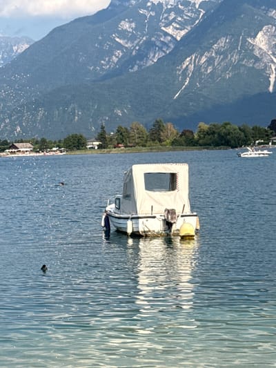 A calm, blue lake reflects the surrounding mountains and a small boat. The boat, moored in the foreground, is white with a tan canopy. A yellow buoy floats beside it. In the distance, a white motorboat carries passengers. The shoreline is lined with trees and buildings. Several birds are visible in the water, one swimming near the boat. The sky is partially cloudy. The scene is set in Calceranica al Lago, Italy.