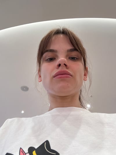 A photograph taken from a low angle shows a fair-skinned woman with brown hair and bangs looking directly at the camera. She is wearing a white T-shirt featuring a cartoon character with a top hat and bowtie. The background appears to be a white ceiling with recessed lighting. The location is Olginskaya, Russia.