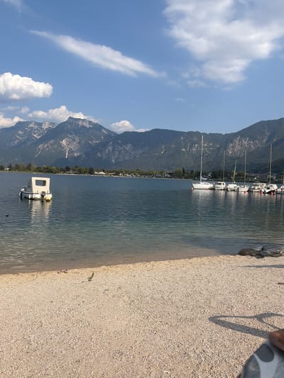 The image depicts a serene lakeside scene in Calceranica al Lago, Italy, under a bright blue sky with scattered white clouds. In the foreground, a sandy beach slopes gently towards the clear, rippling water. A pair of pink flip-flops rests on the sand near the water's edge.
The lake itself is calm, reflecting the sky and the distant mountains. A small, white motorboat with a canvas canopy is moored near the shore on the left. Further out in the lake, a line of white sailboats are docked, their masts tall against the backdrop of rugged, tree-covered mountains. A few buildings and trees are visible along the shoreline in the middle ground. The overall impression is one of tranquility and natural beauty, a typical summer day in a lakeside town.