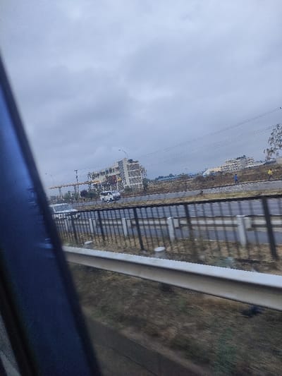 A hazy day with overcast skies. The view is from inside a moving vehicle. In the distance, a multi-story building is under construction. Several vehicles are near it, including a white van and a silver car. Power lines run horizontally across the frame. A guardrail is in the mid-ground. The foreground is blurred, with a blue object partially visible on the left. The scene appears to be an urban or suburban area next to a highway.