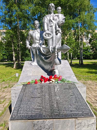 This is a medium-shot, eye-level, outdoor, natural light photograph of a war memorial located in the city of Buy, Russia. The memorial is made of silver-toned metal and depicts three figures on a granite base: a male soldier in uniform, a woman holding a baby, and another male soldier. The woman and the baby are on the right, and the soldiers are on the left. The male soldier in the foreground is seated, and the soldier behind him is standing. The figures are life-sized or larger. There is a wreath of flowers held by the standing soldier. At the base of the memorial, a granite plaque lists names and dates, and a bouquet of red carnations and white tulips is placed on it. The memorial is set against a backdrop of lush green trees and a clear blue sky. The ground around the memorial is covered in green grass, and a paved walkway is visible in the lower right corner. A metal chain is also visible to the left of the memorial, running along the edge of the grass. The overall scene conveys a sense of solemn remembrance and tribute to fallen soldiers.