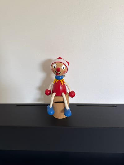 A wooden toy figure, resembling a clown with a red body, blue feet, and a red and white hat, sits on a black surface against a white wall. The figure has a painted face with a red nose, wide eyes, and a pink tongue sticking out. It is wearing a blue collar and a yellow ruff. The figure's arms are attached by white strings to red balls, and its legs are also made of blue balls attached by strings. The toy is positioned centrally in the frame, with the black surface extending horizontally across the bottom of the image. The white wall provides a plain background, with no visible features or decorations. The lighting appears to be natural, casting a soft shadow to the right of the toy. This scene is likely an indoor setting, potentially a home or a shop, and is not specific to any particular location within Drumettaz-Clarafond, France.
