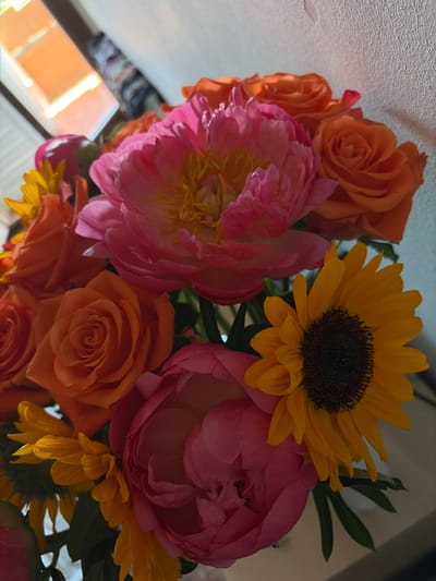 This is a close-up, overhead shot of a vibrant bouquet of flowers. The arrangement features a prominent bright yellow sunflower with a dark center, surrounded by large, lush pink peonies and several orange roses. Small yellow flowers, possibly a variety of aster or daisy, are interspersed among the larger blooms, adding texture and depth. The background is mostly out of focus, showing a textured, light-colored surface and hints of greenery from the bouquet. The lighting suggests it might be daytime, with warm tones illuminating the flowers, particularly the orange roses and the yellow of the sunflower. The overall impression is one of natural beauty and freshness, likely a decorative arrangement rather than a part of a specific event. There is no visible text in the image. Given the context of Riudoms, Spain, the flowers are likely a reflection of local flora or a thoughtful gift.
