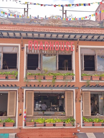 The image shows the facade of a brick building in Gokarneshwar, Nepal. A large red sign above the second-story windows reads "ROADHOUSE". The building has multiple windows and balconies. Prayer flags are strung across the top of the building and extend out of frame. The windows on the second floor are open, revealing interior spaces with plants on the window sills. The windows on the first floor have wooden shutters and more plants on the window sills. People are visible inside some of the ground floor windows, appearing to be staff members. The overall impression is of a restaurant or cafe.