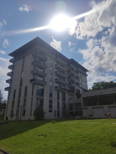 The image shows a large, modern hotel building with a light-colored stone facade and multiple balconies. The building is situated in a green, grassy area with trees visible in the background. The sun is shining brightly in a blue sky with scattered clouds, creating a lens flare effect. Text on the building reads "AMBASSDORI BORJOMI LIKANI". This indicates the location is the Ambassadori Borjomi Likani hotel in Borjomi, Georgia. There are no people or animals visible in the image. The overall scene depicts a sunny day at a resort or hotel.