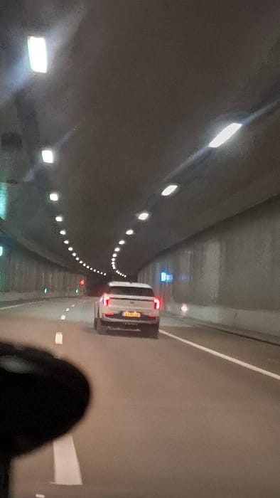A car with illuminated tail lights is driving through a tunnel, its license plate barely visible. The tunnel's interior is illuminated by evenly spaced lights along the curved walls. A dark, blurry object, possibly a person's head, is visible in the lower foreground. The tunnel itself appears to be constructed of concrete. The image is rotated clockwise 90 degrees.