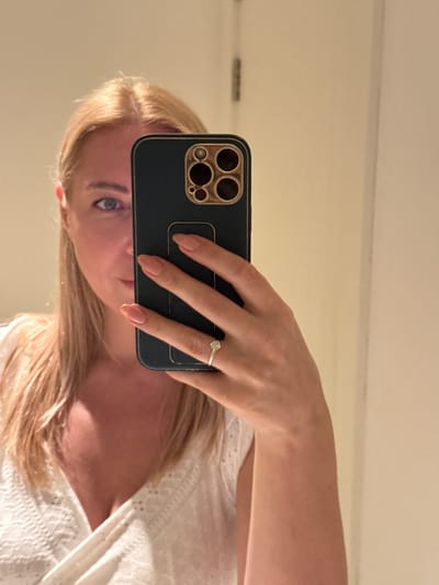 A blond woman is taking a selfie in front of a mirror. She is holding a dark-colored smartphone with a gold-colored camera frame in her right hand, partially covering her face. Her left hand is visible, adorned with a ring. She is wearing a white top. The background is a neutral-colored wall. There is no definitive information in the image to indicate this photo was taken in Ghadeer Al Tayr, United Arab Emirates.