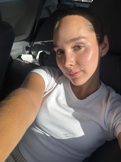 A female individual is observed seated in the rear passenger compartment of a vehicle, actively engaged in capturing a self-portrait. She possesses dark hair, pulled back, revealing her forehead and light brown eyes. Her skin appears moisturized, exhibiting a slight sheen, particularly on the forehead and cheeks, with a subtle rosy tint on the cheekbones. She wears a white, short-sleeved, crew-neck t-shirt and a small, spherical, light-colored stud earring in her visible left earlobe. Her right arm, extended towards the camera, suggests she is holding the imaging device. Strong direct sunlight illuminates the right side of her face, arm, and chest, casting distinct shadows on her shirt and the vehicle's interior. The vehicle's interior features dark, likely leather or synthetic, upholstery on the visible passenger seats and headrests. In the background, on the left side of the rear compartment, a light-colored object, possibly a box or papers, is visible near a dark, indistinct object that may be footwear. The specific location within Ciudad Victoria, Mexico, is not discernible from the visual information provided.