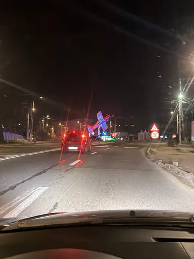 The image depicts an urban street scene at night in Tuzla, Constanța County, Romania, observed from the interior of a vehicle. The perspective is through the windshield, revealing the dashboard and reflections in the foreground. The asphalt road ahead is illuminated by the observer's headlights and various artificial lights, displaying white painted lane markings including a crosswalk on the left.

A red hatchback with illuminated taillights is positioned ahead, displaying a Romanian license plate (CT 77 OHE). To the left of the road, a large decorative light installation, primarily blue and red, shaped like a cross or windmill, is prominently lit. Adjacent to this structure are a circular blue mandatory left-turn sign and an inverted triangular "Yield" sign. On the right side of the road, a circular red-bordered speed limit sign displaying "30" is visible. Further along the right side, beyond the signage, a group of multiple white, vertical structures, resembling religious shrines or monuments, are present.

Utility poles with associated overhead wiring line both sides of the street. A solitary pedestrian figure is discernible on the far right sidewalk, adjacent to a street sign that appears to be a "Stop" sign. The sky is dark.