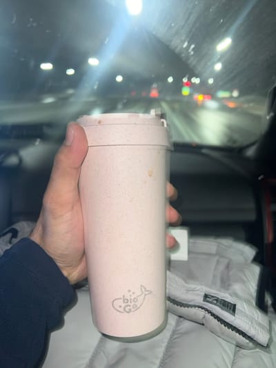 A close-up photograph taken from inside a vehicle, situated in the Town of Fishkill, United States of America, depicts a person holding a travel mug at night. A human left hand, clad in a dark blue long-sleeved garment, firmly grasps a light pink, speckled travel mug. The mug features a grey "bio Go" logo incorporating a whale tail design near its base and exhibits minor brown stains on its upper body and lid. To the right of the mug, a folded white quilted jacket or similar garment is visible on a seat. Through the vehicle's windshield, an exterior night scene unfolds, characterized by blurred white overhead lights, red and yellow vehicle tail lights, and reflections on a wet road surface, suggesting movement or a recent stop during damp conditions. The interior of the vehicle, including parts of the dashboard, is faintly discernible in the lower background.
