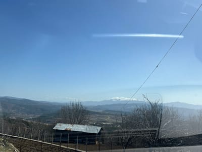 The image presents a panoramic view of a mountainous rural landscape near Krushevo, Bulgaria, under a clear blue sky. A distinct horizontal light reflection is visible across the upper right portion of the sky, bisected by a thin, dark diagonal wire extending from the upper edge towards the lower right. In the midground, a single, small, rectangular structure with dark walls and a rusty corrugated metal roof is situated slightly to the left of the image center. Immediately to its left, a fenced plot of tilled or planted dark earth is visible, secured by dark vertical posts. Several bare deciduous trees are scattered across the midground. The terrain slopes downward from the foreground into a valley containing a visible settlement with scattered buildings. Distant rolling hills and mountains, including a range with prominent snow-capped peaks, complete the background. No individuals or specific actions are depicted within the scene.