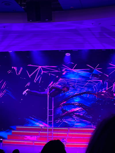 In a dimly lit, vibrant scene, a man hangs from a horizontal bar, his legs dangling. He wears a dark outfit and white shoes. The backdrop features geometric neon lights, a tree-like structure, and vertical strips of bright lights. The lighting casts a strong purple hue, particularly prominent on a large, vertical structure to the left. A partial silhouette of an audience member is visible on the right.