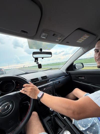The image displays the interior of a moving vehicle from a low, front passenger perspective, looking towards the driver and the road ahead. The scene is identified as being in Bragadiru, Romania.
A male is in the driver's seat of a left-hand drive vehicle, with his right hand gripping the black steering wheel that features red stitching and a central Mazda logo. An Apple Watch with a dark band is worn on his left wrist, displaying a red and pink pattern. He has short dark hair and stubble, and the right side of his face, showing a slight smile, is visible as he looks towards the right. He is wearing a white short-sleeved t-shirt and light-colored, possibly bleached, denim shorts. His bare forearms and legs are visible.
The dashboard is dark-colored. The speedometer, marked up to 220 km/h, displays an illuminated orange warning light. The center console includes a radio/CD player unit and climate controls. A manual gear stick and an emergency brake lever are visible to the right of the driver's seat.
Through the windshield, a multi-lane asphalt highway extends into the distance under a partly cloudy sky. Another dark vehicle is observed in the left lane ahead. Green agricultural fields flank both sides of the highway, with utility poles or towers visible on the distant horizon. A grey, angular structure, possibly a sound barrier or ramp, is visible on the right side of the highway. A rearview mirror is mounted on the windshield, along with a black magnetic phone holder. The vehicle's interior roof lining and sun visors are grey; the passenger-side sun visor features a yellow warning label. An overhead interior light fixture is visible on the passenger side. The passenger door panel is dark with a silver handle.