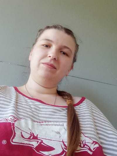 A young woman with light skin, brown hair styled in a braid, and dark eyes looks up at the camera. She is wearing a grey and white striped shirt with a red neckline and a graphic print on the front. The background is a plain, light green wall with a horizontal line running across it. The image appears to be taken indoors, possibly in a residential setting. There is no indication of the location being Kitskany, Moldova.