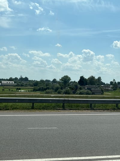 The image depicts a rural landscape with a clear blue sky filled with scattered white clouds. In the foreground, there is a section of asphalt road with white lane markings, bordered by a metal guardrail and grassy verges. Beyond the guardrail, a green field stretches towards a cluster of buildings, suggesting a small village or hamlet. The buildings appear to be residential, with pitched roofs and varying architectural styles, surrounded by trees. The overall impression is that of a tranquil, sunlit day in a countryside setting, possibly near Мастоцкі сельскі Савет, Belarus. No people or animals are visible in the frame.