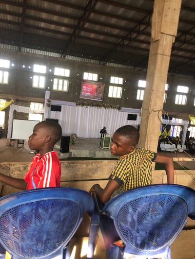 A medium, angled shot inside what appears to be a large hall or church in Nteje, Nigeria. Natural light streams through numerous windows, illuminating the interior. The hall has a high ceiling with exposed beams and a metallic roof. At the far end of the hall, a stage is decorated with white fabric, flowers, and a large banner. A man stands at the podium on the stage.
In the foreground, two young Black boys are positioned near rows of blue plastic chairs. One boy wears a yellow and black checkered shirt, and the other wears a red shirt with white stripes. They both appear to be looking downward, possibly observing something on the floor or ground. The blue chairs are stacked close to the camera, partly obscuring the view of the boys.