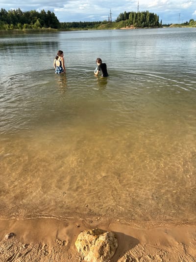 In the city of Zamost'e, Russia, two individuals are wading in a shallow body of water. The person on the left, facing away from the camera and looking towards the right, wears a dark swimsuit with a floral pattern. The person on the right, also facing away and slightly to the left, is clad in a black wet suit or dark-colored long-sleeved garment. The water is clear enough to reveal a sandy bottom. The background features a dense line of trees and a hazy sky with clouds and power lines. The foreground shows a sandy shore with a large, light-colored rock.