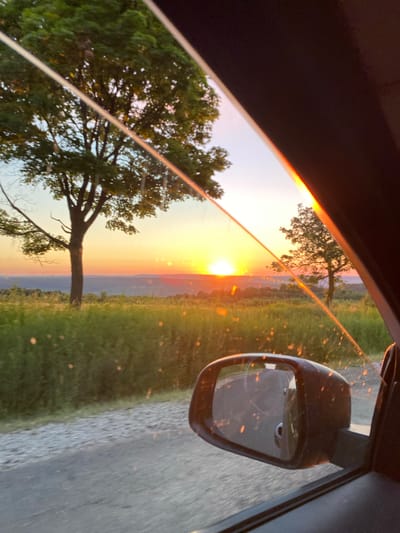 The image captures a sunset viewed from inside a vehicle, likely a car, looking through the windshield and side window. The setting sun paints the sky in vibrant hues of orange and yellow, casting a warm glow over a rural landscape. In the foreground, the side mirror of the car is visible, reflecting a portion of the road and the surrounding environment. A gravel road runs alongside a grassy field, and trees dot the landscape. The overall scene evokes a sense of peaceful travel during a beautiful sunset. The location is identified as Vulcăneşti, Moldova.