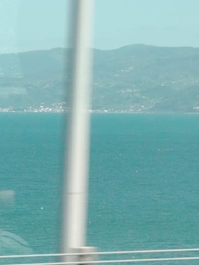 The image captures a hazy daytime view of a wide body of water, likely a sea or a large bay, with a mountainous coastline in the distance. In the immediate foreground, a prominent vertical, light-colored pole runs from the top to the bottom of the frame, slightly off-center to the right, partially obscuring the scene. Two thin, white horizontal rails are also visible at the very bottom of the frame, suggesting the photograph was taken from behind a barrier or through a window.
The water, which occupies the majority of the midground, is a striking shade of turquoise or teal, exhibiting subtle textural variations that indicate a slightly choppy surface. On the distant coastline, a scattering of small, light-colored buildings forms a town or settlement along the water's edge, nestled against the base of the mountains. The mountains themselves are muted in color, appearing as soft blue-grey forms under a light blue, somewhat hazy sky.
There are no discernible people or explicit activities taking place, as the image focuses on the landscape. The overall atmosphere is serene but indistinct due to the haze. No visible text is present in the image. The scene is consistent with a coastal view, potentially overlooking a body of water such as the Gulf of İzmit from Dilovası, Turkey, given the provided location context.