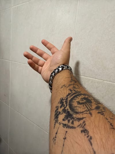 A person's forearm, adorned with a tribal-style tattoo and a silver link bracelet, is positioned against a tiled wall. The arm is bent, with the hand open and fingers extended upwards. The tiled wall, a light beige color, features distinct grout lines creating a grid pattern. The lighting suggests an indoor environment. The image is focused on the forearm and hand, with the background out of sharp focus. There are no discernible geographical markers to indicate a specific location in Torremolinos, Spain.