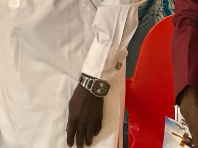 The image shows a close-up of a person's arm wearing a wristwatch. The person is wearing a white long-sleeved shirt with a cufflink on the sleeve. The wristwatch has a metallic band and a dark face with two circular elements. A hand with dark skin is visible, with the watch on the wrist. In the background, a bright red object, possibly a chair, is visible. To the left of the red object, a white garment is seen, which appears to be part of the same shirt as the sleeve. Further to the left, a glimpse of a patterned fabric is visible. To the right of the red object, a portion of a dark red garment is seen. At the bottom right, a part of a picture or print is visible, showing a cross and some sky. The overall setting suggests an indoor environment. There are no explicit cues about the time of day or weather, nor any visible text. The activity taking place is not evident, other than the person wearing the watch. The image appears to be taken in Kufana, Nigeria, as per the location context.