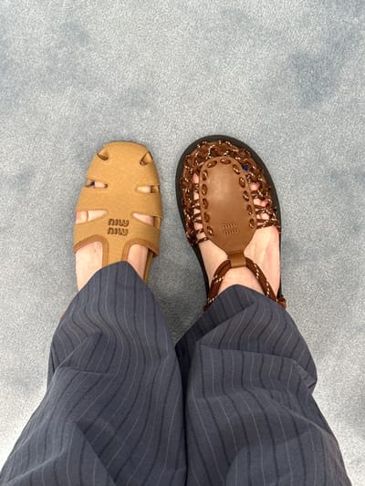 The image shows a person wearing mismatched Miu Miu sandals while standing on a light blue carpet or rug. The sandal on the left foot is brown and beige with a woven strap design, while the sandal on the right foot is light brown with cut-out details. The person is wearing dark blue pinstripe pants. There is no indication of the image being taken in Costa Mesa, United States of America.