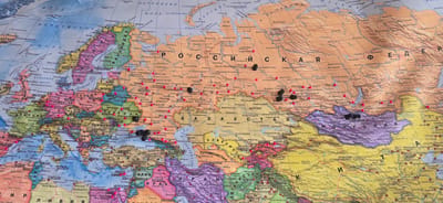 The image is a close-up of a world map, specifically focusing on Europe and parts of Asia. The map is a flat, two-dimensional representation with political boundaries, city names, bodies of water, and mountain ranges depicted in various colors. Overlaid on the map are numerous black and red push pins, marking various locations. Some pins are clustered together, while others are spread out. The pins suggest points of interest, travel destinations, or perhaps a record of places visited or to be visited.
The map itself is detailed, with labels for countries like Russia, Ukraine, Poland, Germany, France, Spain, Italy, and Scandinavian countries. Major cities are clearly marked with dots and names, such as Moscow, Berlin, Paris, London, Rome, and Kyiv. Bodies of water like the Mediterranean Sea, Black Sea, Baltic Sea, and Atlantic Ocean are also visible.
The push pins are the primary objects of focus, drawing attention to specific points on the map. The black pins appear to be more numerous in certain regions, particularly across European Russia and parts of Siberia, as well as in Western Europe. Red pins are also scattered across the map, with a notable concentration in some areas of Eastern Europe and Central Asia.
The scene is static, as it's a photograph of a map. There are no people or active events depicted, only the representation of geographical locations and the markers on them. The lighting suggests it was taken in a well-lit indoor environment, likely to capture the details of the map clearly. The time of day and weather are not discernible from the image.
The location context provided, "Gostagayevskaya, Russia," is a specific settlement in Russia. While the map itself covers a vast area, this specific location would be a small dot on the map, likely within the Krasnodar Krai region of southern Russia. The pins might be related to this location, perhaps indicating travel to or from it, or marking it as a point of significance. The map is a tool for understanding geography and planning journeys, and the pins are a personal annotation on that representation.