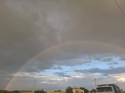 A rainbow arches across a cloudy sky over Chorna, Ukraine. The foreground is dominated by a landscape of trees, a small building, a utility pole, and a truck. The sky is filled with a mix of dark storm clouds and lighter, scattered clouds. The rainbow, a semi-circular arc of colors, is visible against the darker clouds. In the lower right quadrant of the image, the front of a white and blue truck is visible. To the left of the truck, a small, light-colored building with a red door is partially obscured by trees. A utility pole stands near the truck. The ground appears to be covered in grass and scattered foliage.
