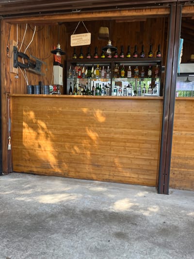 The image depicts a partially open outdoor bar, possibly in Valchi Dol, Bulgaria, given the language of the sign visible. The bar's interior has wooden walls and a shelf holding an assortment of bottled alcoholic beverages. A sign, in what appears to be Bulgarian Cyrillic, is attached to the bar's interior. Below the shelves, there are electrical wires, a light fixture, and what seems to be a TV mounting bracket. On the right side, a large section of the bar is a sliding wooden panel, partially covering the interior. The exterior floor is a concrete surface. No people are visible in the frame.