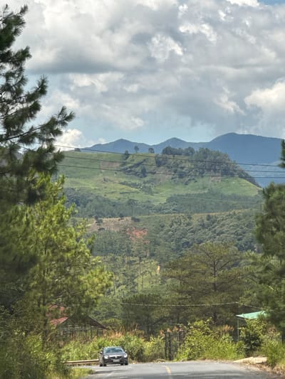The image captures a scenic view of a mountainous landscape in Tu Póh, Vietnam. In the foreground, a paved road curves to the right, with a dark-colored sedan driving towards the viewer. The road is bordered by lush green vegetation and tall pine trees on either side. In the middle ground, rolling hills covered in various shades of green, from dense forests to terraced fields, rise towards the horizon. Some areas of exposed red earth suggest agricultural activity or land clearing. Power lines are visible crisscrossing the hills. In the background, layers of blue-hued mountains fade into the distance under a sky filled with dramatic, light-colored clouds. A small, reddish-roofed building is partially visible amidst the foliage on the left side of the road. The overall impression is one of natural beauty and rural tranquility.