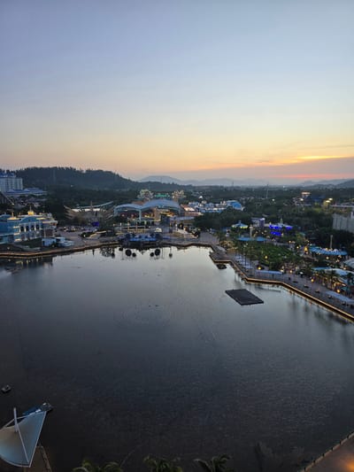 This is a high-angle, wide shot of a waterfront amusement park in Li'an, China, during twilight. The sky transitions from a pale blue at the top to a warm orange and yellow near the horizon, indicating either sunrise or sunset. The park features a large, dark body of water in the foreground, with the shore lined by a promenade illuminated with string lights.
On the left side of the water, a light blue, multi-story building with white trim is visible. Across the water, the amusement park complex is illuminated with colorful lights. Various attractions, including what appear to be roller coasters, a Ferris wheel, and stage structures, are present. Green trees and lush landscaping are interspersed throughout the park. In the distance, hills covered in trees form the backdrop, with power lines and distant structures visible.
Several small, dark objects, possibly boats or rafts, are scattered across the surface of the water. A rectangular floating platform is positioned near the center of the water. There are also clusters of small, light-colored dots in a circular formation on the water's surface, likely part of a fountain or lighting display. A few people are visible walking along the illuminated promenade. The overall scene depicts a vibrant, yet tranquil, amusement park setting as daylight fades.