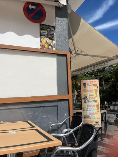 The image shows an outdoor seating area of a cafe in Agüimes, Spain, at the location of (latitude: 27.907, longitude: -15.447). There are multiple brown wooden tables with black trim, and several black chairs with silver frames around them. There is a large white awning above the tables, providing shade. Behind the tables are several large glass windows, one of which displays a menu in Spanish. To the left of the window, a smaller white board with a brown wooden frame is mounted to the wall. On the wall to the left of the window is a colorful advertisement for a cafe, and above it is a no-entry sign.  A cafe menu is also displayed in the image, with a variety of smoothies, milkshakes, and desserts listed on it.  A black metal stand holding the menu sits on the floor, and behind it, the blurred background suggests a patio or outdoor area with plants and a stone wall.  A table with a single chair is visible in the background, indicating other cafe customers.  The tables are empty, and there are no people visible in the image. The sky is blue with a few clouds in the image.