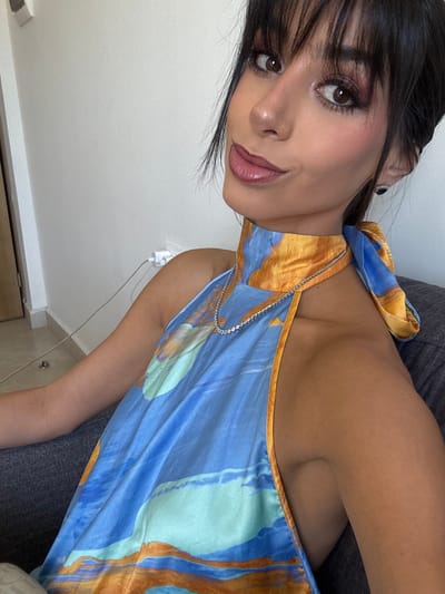 A woman is shown at an upward angle. She has dark hair with bangs and is wearing makeup, including lipstick and eyeshadow. Her skin tone is medium. She wears a halter-style top with a blue and orange pattern, and a delicate silver chain rests on her neck. She is resting on a grey surface. A white wall is visible behind her, with a white electrical outlet extending from it. The room appears to be indoors.