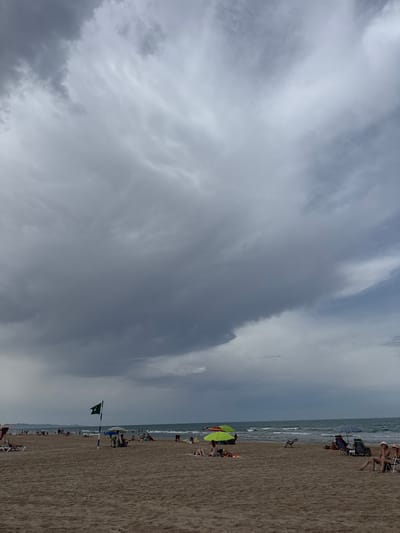 This image captures a scene on the beach at Grau i Platja, Spain, under a cloudy sky. The sand is a light brown, and the sea, with moderate waves, stretches to the horizon. The sky is dominated by large, dark gray clouds, with lighter patches and streaks of white cloud visible.
Several people are present on the beach. In the foreground, there are beach chairs and umbrellas set up. On the left side of the image, two people are seated in lounge chairs. In the middle ground, a green flag with a white symbol is planted on a pole. Further back, more beachgoers are scattered, some sitting under umbrellas, others lying on lounge chairs or towels. People are also visible in the water, engaged in various beach activities. The background shows a hazy coastline with what appears to be buildings. The overall impression is of a somewhat overcast but active beach day.