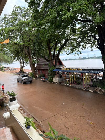 This image captures a waterfront scene in Thakhek, Laos. A gray pickup truck is parked on a wet, reddish-brown asphalt road. To the left of the truck, a person wearing a red shirt and dark shorts stands by the side of the road, holding a smartphone. Further left, a decorative yellow bird sculpture is partially visible.
Dominating the right side of the image is a rustic, wooden building with a red exterior and a brown, tiled roof, which appears to be part of a restaurant or cafe. In front of this building is an outdoor seating area with tables and chairs under a blue-roofed canopy, decorated with yellow and pink floral garlands. A prominent sign above the canopy reads "Beerlao Le Bouton D'Or". A large, stone oven is visible near the building. A scooter is parked near the entrance of the establishment.
The scene overlooks a wide, calm river. The riverbank in the mid-ground shows some construction or landscaping work with piles of dirt and debris. In the distance, across the river, is the skyline of a city, featuring buildings and a Ferris wheel. The sky is overcast, with patches of blue showing through the clouds. The overall impression is of a tropical, slightly humid environment, likely during or after rainfall, given the wetness of the road. Lush green trees with dense foliage are abundant throughout the scene, framing the view of the river and the city beyond. White railings and potted plants are visible in the foreground, suggesting a residential or commercial structure.