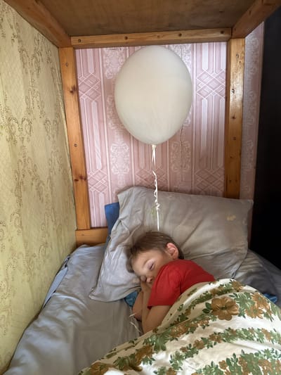 A child in a red shirt is lying on a bed under a floral-patterned blanket, resting on a light grey pillow. A white balloon is tied to the bed frame with a white string. The bed is positioned against a wall with pink and white wallpaper. The image appears to be taken from above, looking down on the scene. The location is not discernible from the image.