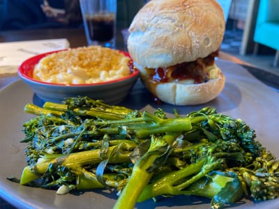 The image presents a plate of food featuring a burger, mac and cheese, and broccolini. The burger is a single patty topped with what appears to be barbecue sauce, served on a soft bun. The mac and cheese is topped with breadcrumbs and served in a red-rimmed bowl. A portion of broccolini, sauteed with garlic, completes the meal. A glass of dark liquid is positioned in the background. The food rests on a gray plate. The scene lacks specific indicators to pinpoint the location within Elmbridge, United Kingdom.